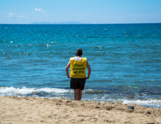 Una ragazza sulla spiaggia che guarda il mare con una pettorina con scritto "stiamo lavorando per noi"