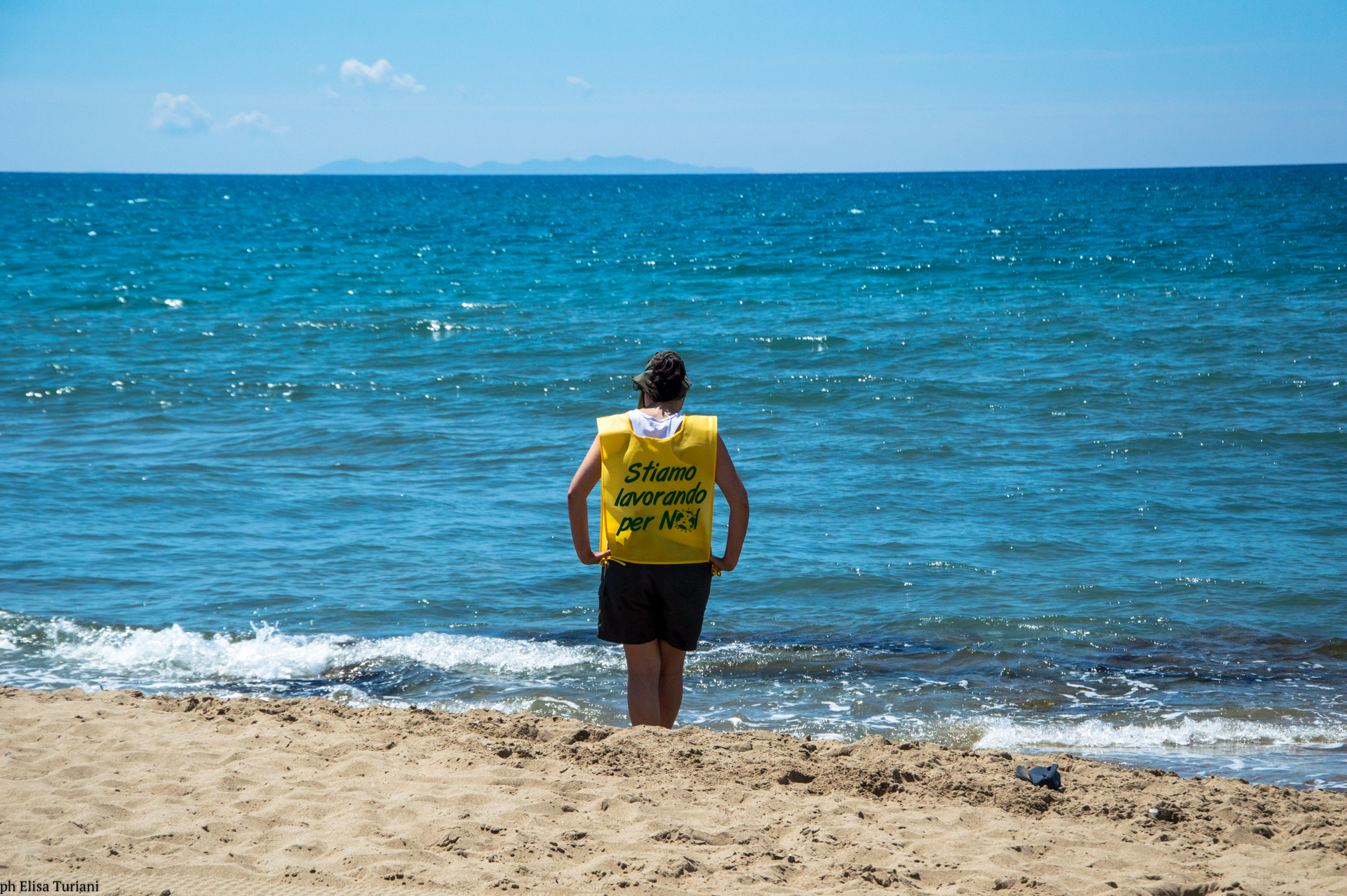 Una ragazza sulla spiaggia che guarda il mare con una pettorina con scritto "stiamo lavorando per noi"