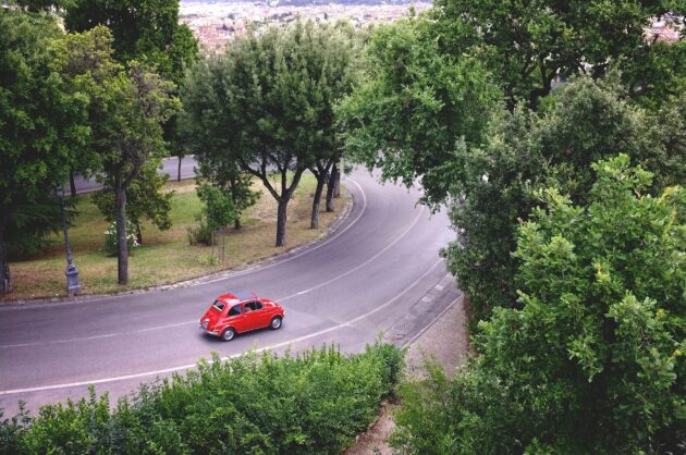 Una cinquecento rossa che passa su un viale alberato cittadino